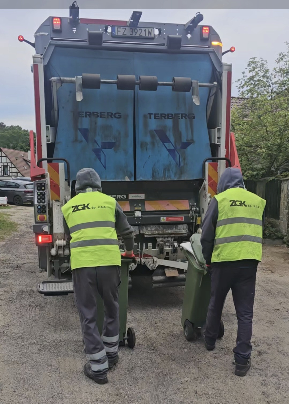 Dwaj panowie w kamizelkach odblaskowych z napisem ZGK odbierają odpady. Na zdjęciu znajduje się także śmieciarka.