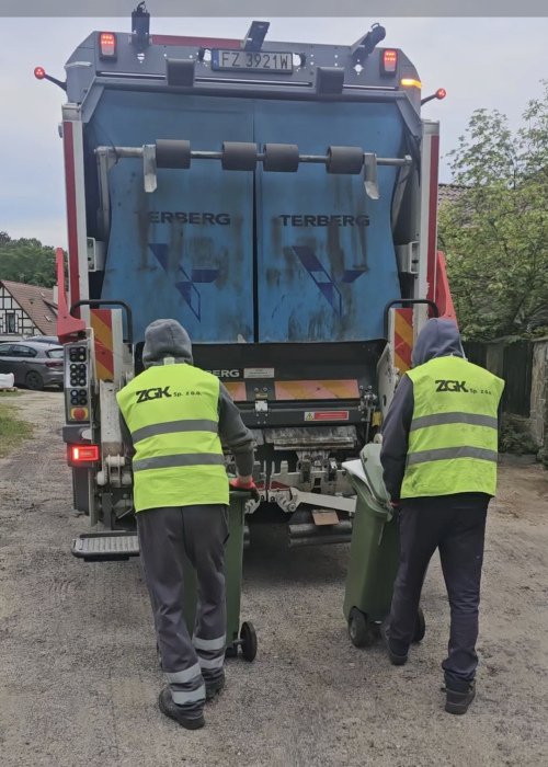 Dwaj panowie w kamizelkach odblaskowych z napisem ZGK odbierają odpady. Na zdjęciu znajduje się także śmieciarka.
